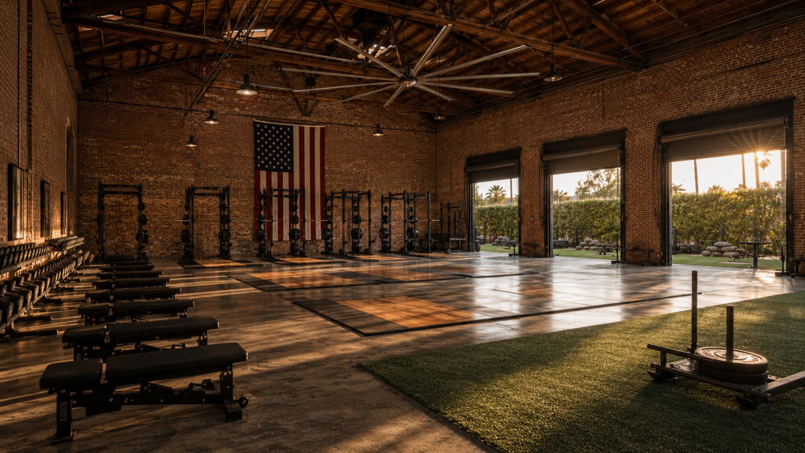 The strength floor at golden hour — brick warehouse, American flag, roll-up doors open to the outdoor strip.